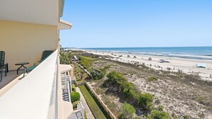 Un balcon doté de mobilier d’extérieur, donnant sur une plage délimitée par une clôture et où se trouvent des personnes se promenant sur le sable.