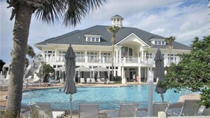 Un resort con una piscina grande, sillas de descanso, sombrillas y un edificio de dos pisos con balcón.