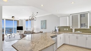 A modern kitchen with granite countertops, white cabinetry, and a large island. There is a dining area with a view of the ocean and a ceiling fan. - Gulf Front Beach Club - Renovated Corner Condo, Direct Gulf Views! Pool, WIFI (Gulf Shores)