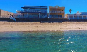 Une propriété située en bord de mer, offrant une vue dégagée sur l’océan, une plage de sable et un immeuble à plusieurs étages doté de balcons.