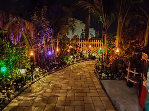 Un chemin pavé, bordé de luminaires décoratifs et de plantes, mène à un bâtiment blanc doté d’une clôture en treillis.