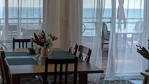 A dining area with a table set for four, surrounded by chairs, with a view of the sea through sheer curtains.