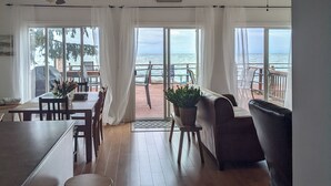 A spacious living area with a large window overlooking the sea, a ceiling fan, and a dining area with a table and chairs.