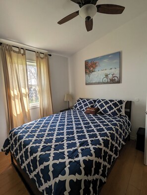 A bedroom with a bed, a ceiling fan, a window with curtains, a painting on the wall, and a nightstand. - Stunning Lake Erie Water Views from Every Window with Expansive Patio Deck
(Leamington)