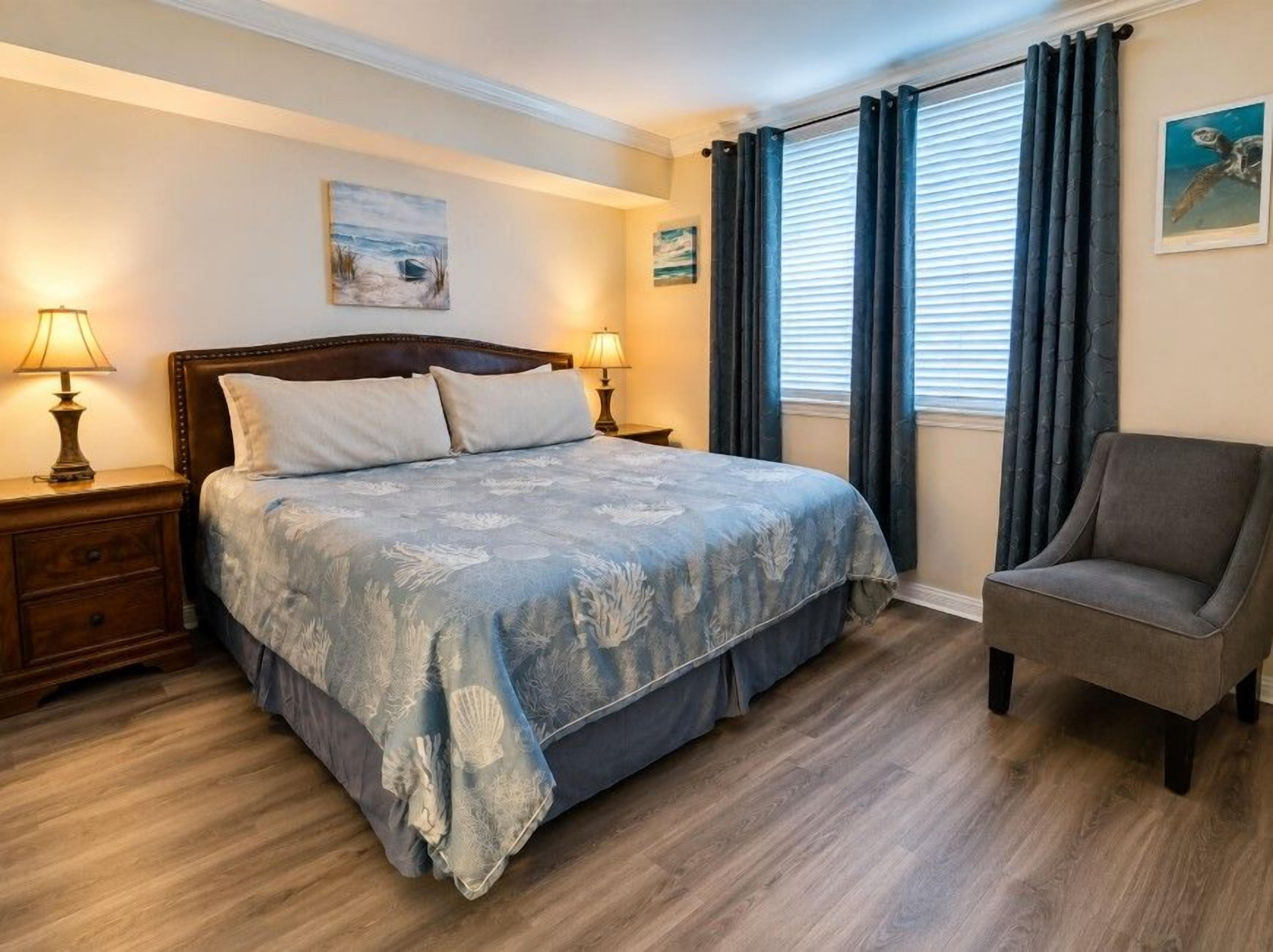 Un dormitorio con cama, mesitas de noche, una silla y un ventanal con cortinas.
