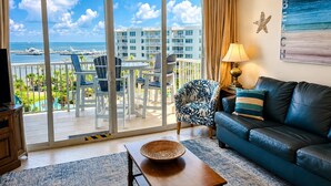 A living room with a sofa, a table, and a chair, a balcony with a view of the sea and buildings, and a starfish decoration on the wall.
