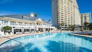 Une grande piscine entourée de chaises longues et de parasols, elle-même bordée par un bâtiment d’hôtel à plusieurs étages doté de balcons.