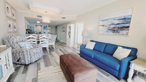 Un salon avec un canapé bleu, un pouf à motifs et des meubles blancs. Au fond, on aperçoit une cuisine avec des armoires blanches et un coin repas.