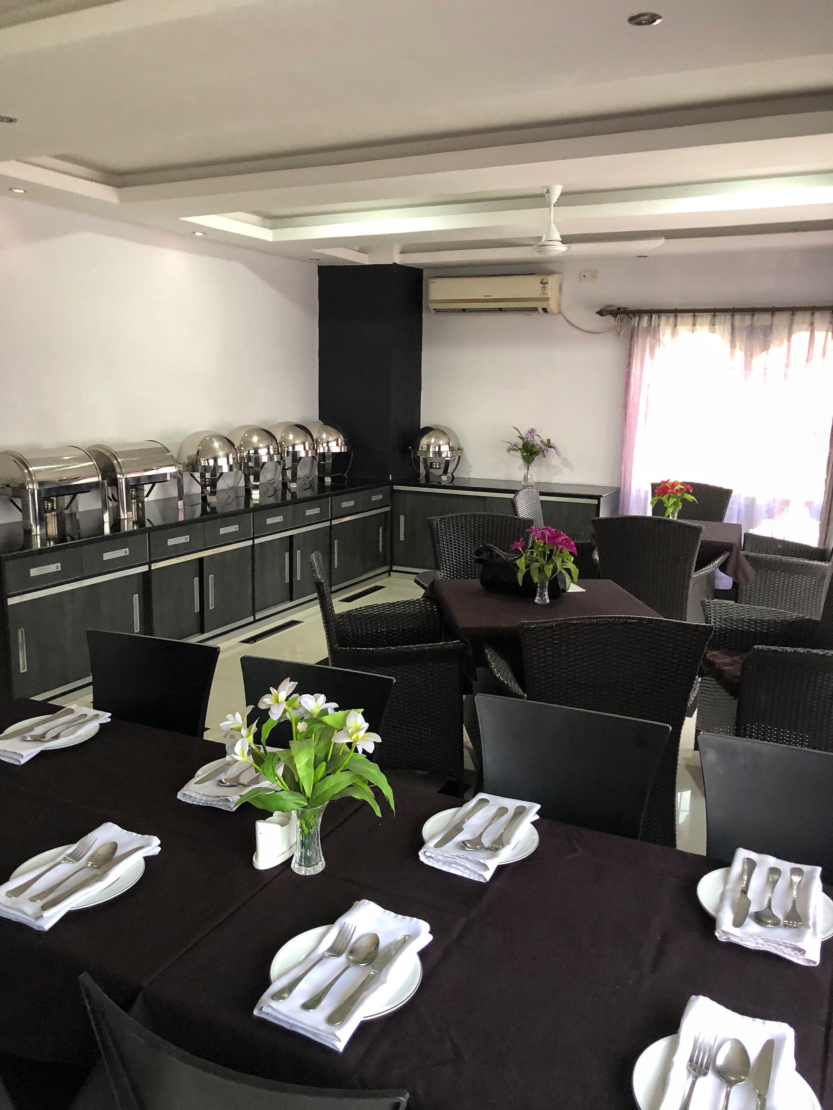 A dining area with a long table set for a meal, surrounded by chairs. There are kitchen cabinets and a window with curtains.