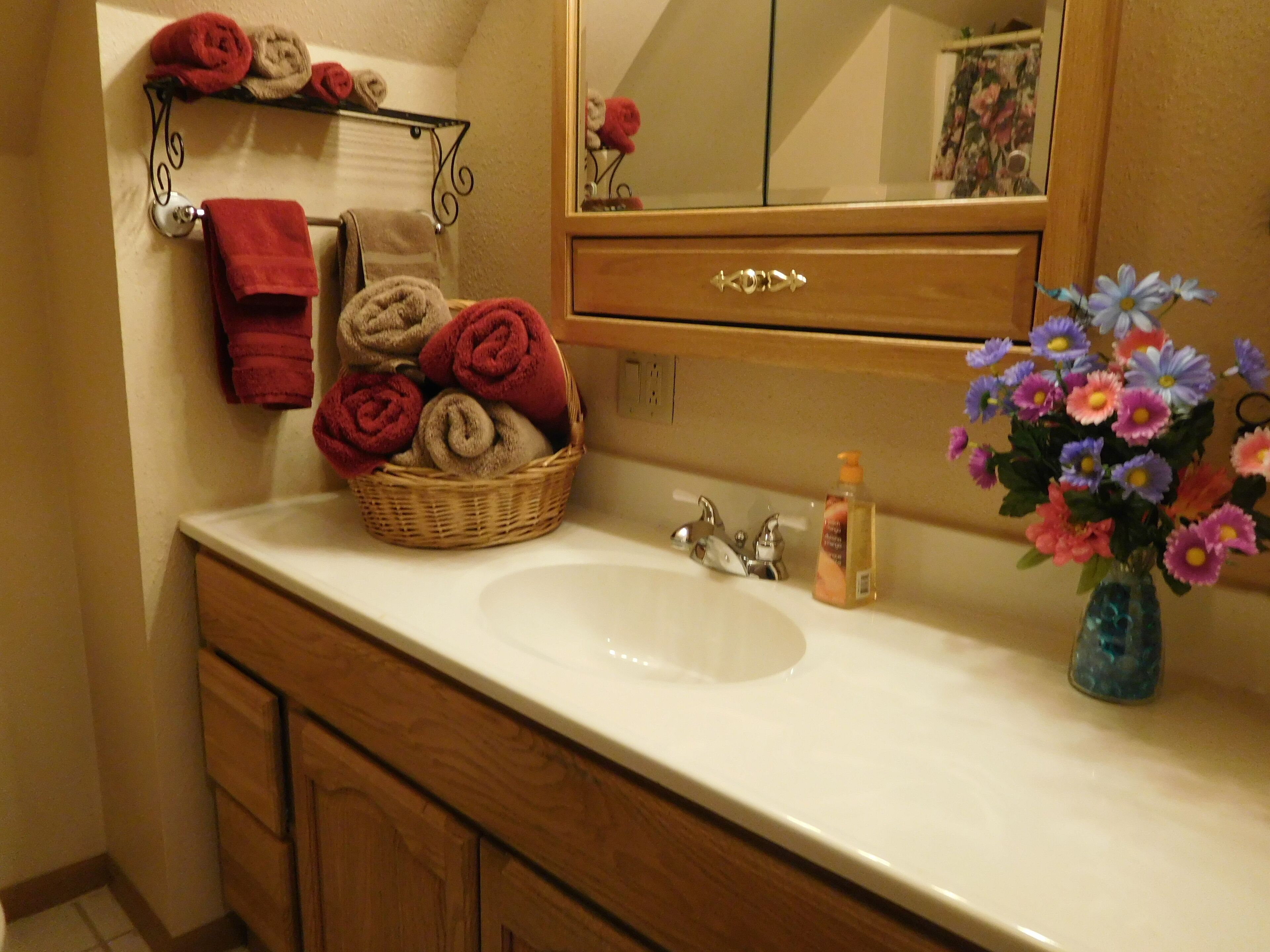 A bathroom with a white sink, wooden vanity, a mirror, a towel rack with rolled towels, a wicker basket with towels, and a vase with flowers.