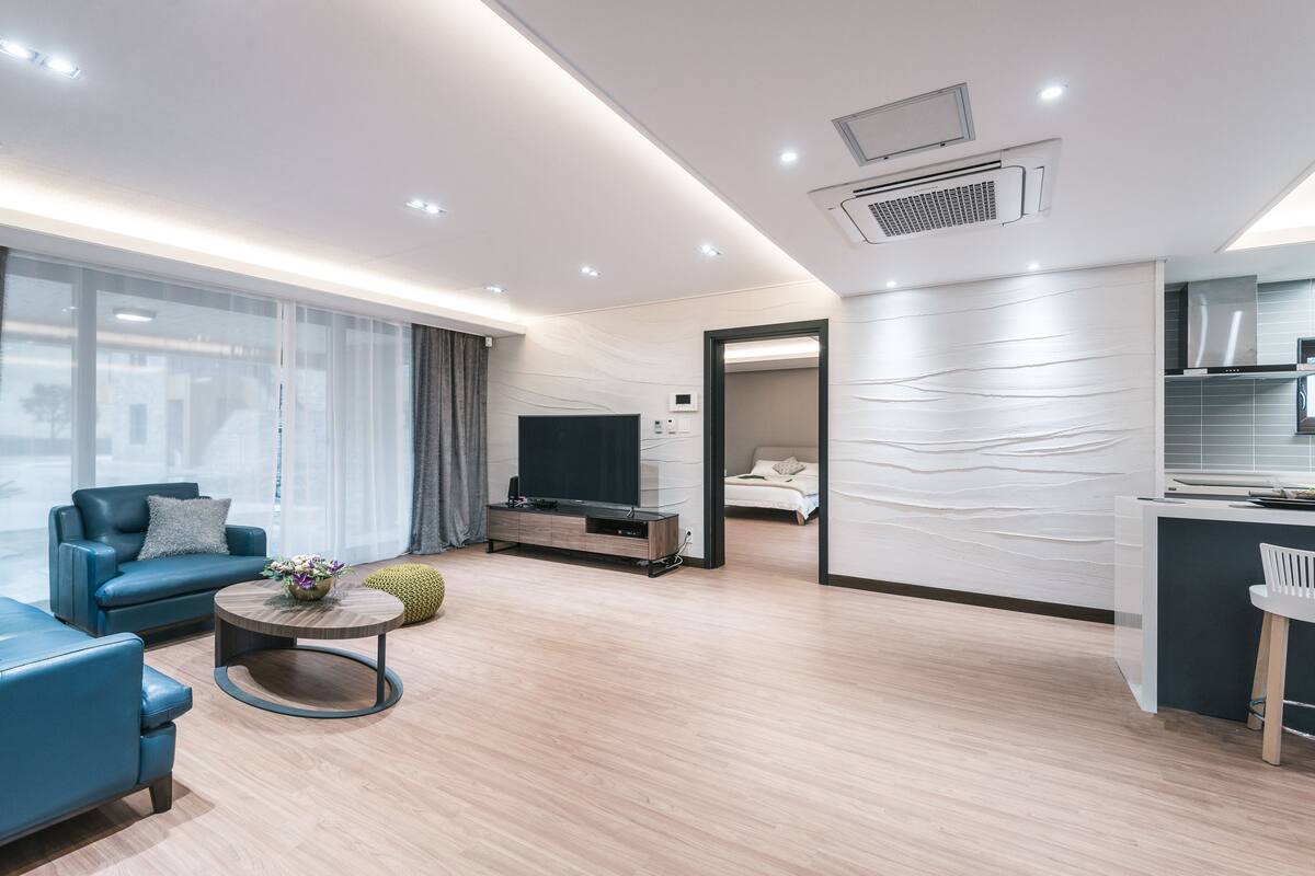 Un salon moderne avec un canapé, une table basse et un téléviseur. Il y a un coin cuisine avec un comptoir et une chaise. La pièce est dotée d’un plancher en bois et d’une grande fenêtre avec des rideaux.
