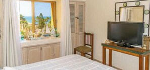 Ein Schlafzimmer mit Bett, Fernseher auf einem Schrank, einem Sessel, einem Fenster mit Blick auf Palmen und das Meer sowie einem Spiegel an der Wand.