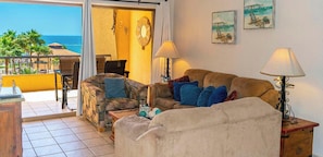 Ein Wohnzimmer mit einer Couch, Sesseln und einem Couchtisch. Es gibt zwei Lampen, ein Gemälde und ein Schild mit der Aufschrift 'Welcome' an der Wand.