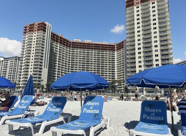 A beach with lounge chairs labeled 'Shores of Panama' and a high-rise hotel in the background. - Gulf-Front Corner 3BR w/ Wrap Balcony + Sunset Views | Beach service included (Panama City Beach)
