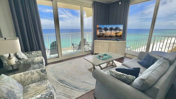 Ein Wohnzimmer mit einer Couch, Couchtisch und Fernseher, mit Blick auf einen Strand mit Palmen und einem klaren Himmel.