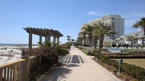 Un sentiero conduce a una spiaggia con un pergolato, palme e un albergo sullo sfondo.