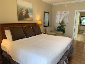 A large bed with white linens and multiple pillows, a wooden headboard, a side table with a lamp, a chair, and a potted plant.