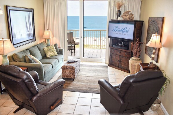 A living room with a sofa, two leather chairs, a television, and a view of the beach.