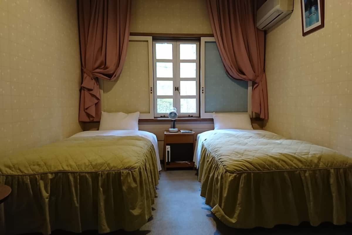 Quarto com duas camas, mesa de cabeceira com abajur, janela com cortinas e ar condicionado.