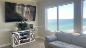 Een woonkamer met een grote flatscreen televisie aan de muur, een witte console met een plant en een boek, een grijze bank en een groot raam met uitzicht op de oceaan.