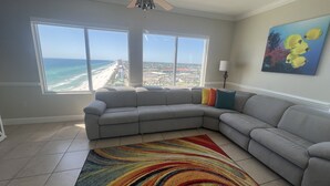 Een woonkamer met een groot raam dat uitkijkt op een strand en stadsbeeld, een grijze hoekbank, een kleurrijk vloerkleed en een schilderij van vissen.