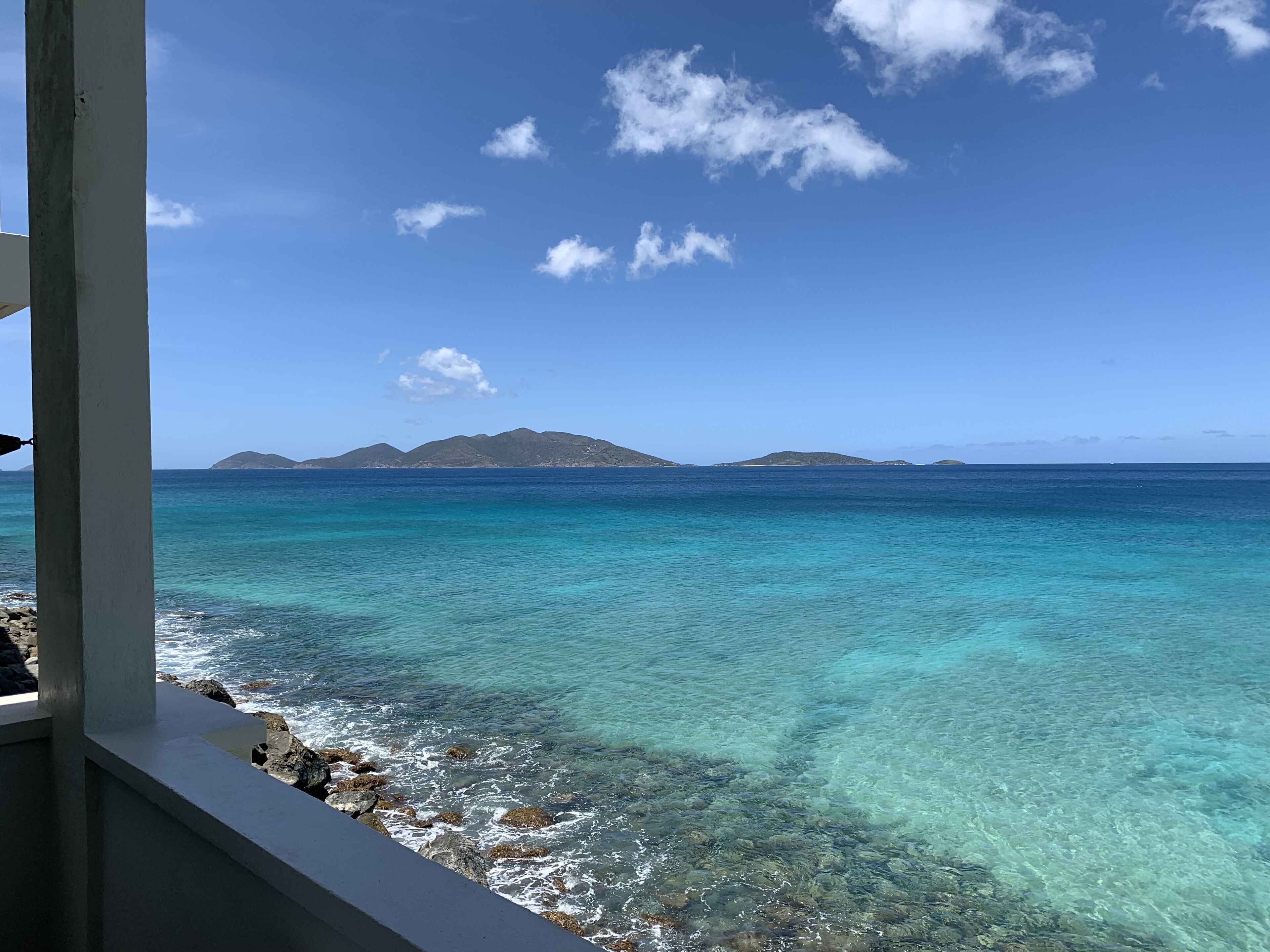 A coastal view with clear turquoise waters, rocky shoreline, and distant islands under a clear blue sky.