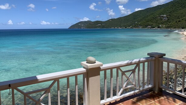 A clear view of a turquoise sea from a balcony with a white railing, a rocky shoreline, and a green hill with buildings in the background. - CASA CARIBE OCEANFRONT TWO STORY LOFT VILLA, Air Conditioning, Beautiful Views. (Little Apple Bay)