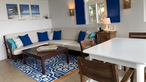 A modern living room with a white sofa, blue cushions, a wooden coffee table, and a dining area with a white table and wooden chairs. - CASA CARIBE OCEANFRONT TWO STORY LOFT VILLA, Air Conditioning, Beautiful Views. (Little Apple Bay)