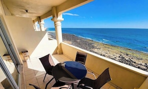 En balkon med et bord og stole, der ud til en stenstrand og havet.