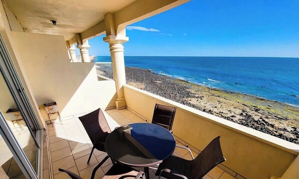 A balcony with a table and chairs, overlooking a rocky beach and the ocean.