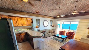 A kitchen with wooden cabinets, a tiled backsplash, and a central island. There is a dining area with a sofa and a coffee table. The room has a brick ceiling and large windows. - Family Condo Sea View Pool | 3bdr Sleeps 7 (Penasco)