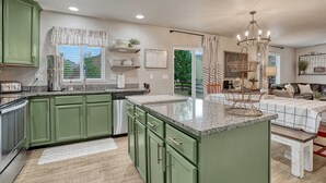 Une cuisine moderne avec des meubles de couleur verte, un plan de travail en granit et un coin repas agrémenté d’un lustre.