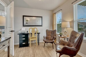 Une pièce comprenant un bureau, deux fauteuils en cuir, une petite table avec une lampe, un miroir au mur et une fenêtre avec des stores.