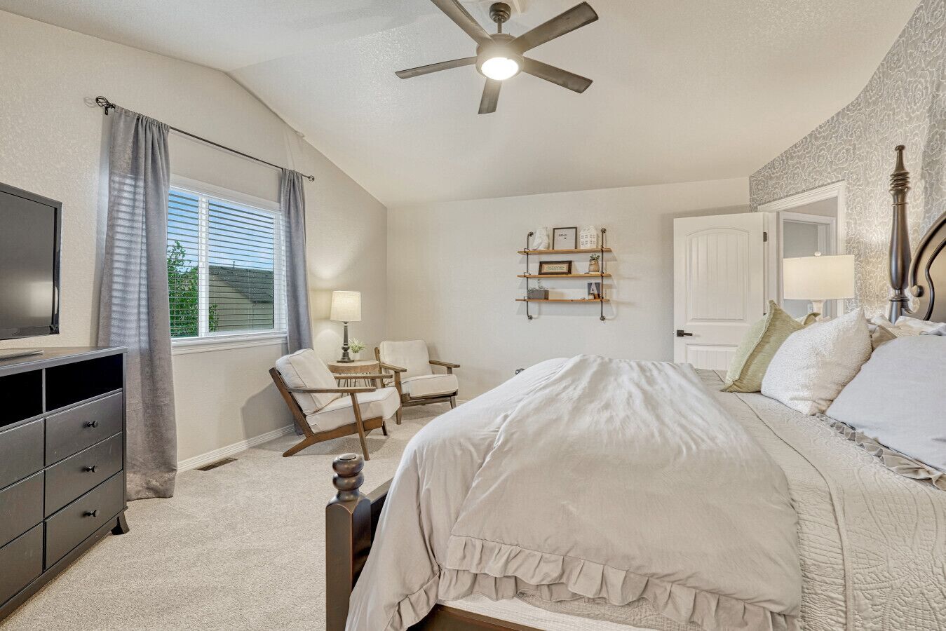 A bedroom with a bed, two chairs, a desk, a TV, and a ceiling fan.