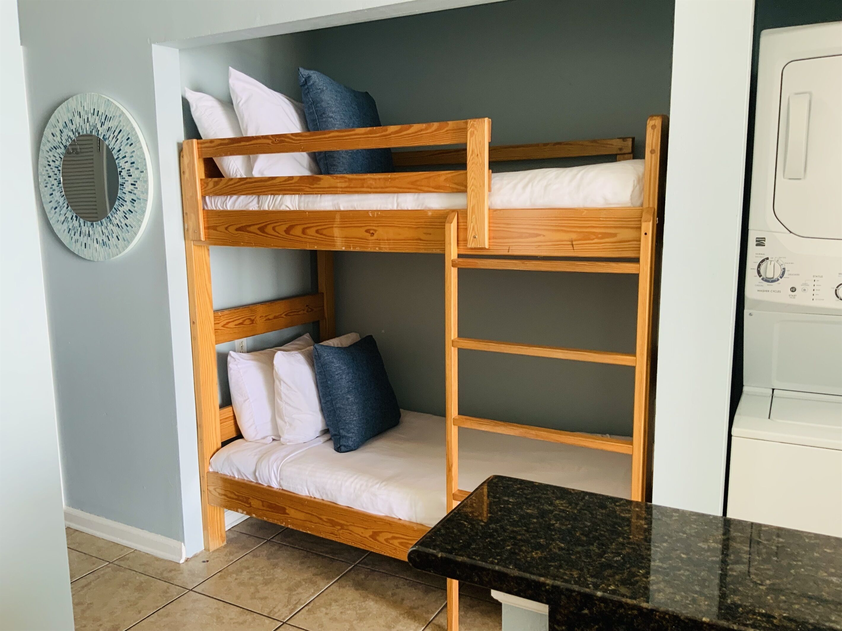 Un lit superposé avec des oreillers, un lave-linge et un miroir fixé au mur, dans une pièce dont le sol est carrelé.