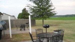 An outdoor patio with a round metal table and chairs, a barbecue grill, and a white fence in the background.