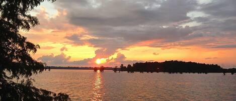 A sunset over a calm body of water with silhouettes of trees and a distant shoreline.
