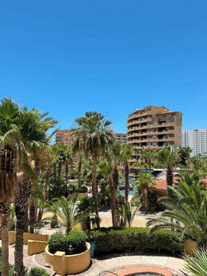 A resort with palm trees, a pool, and a large building in the background. - Bella Sirena B-202 2BR/2BA Upper Floor | Ocean Views & Luxury Sandy Beach Escape (Puerto Penasco)