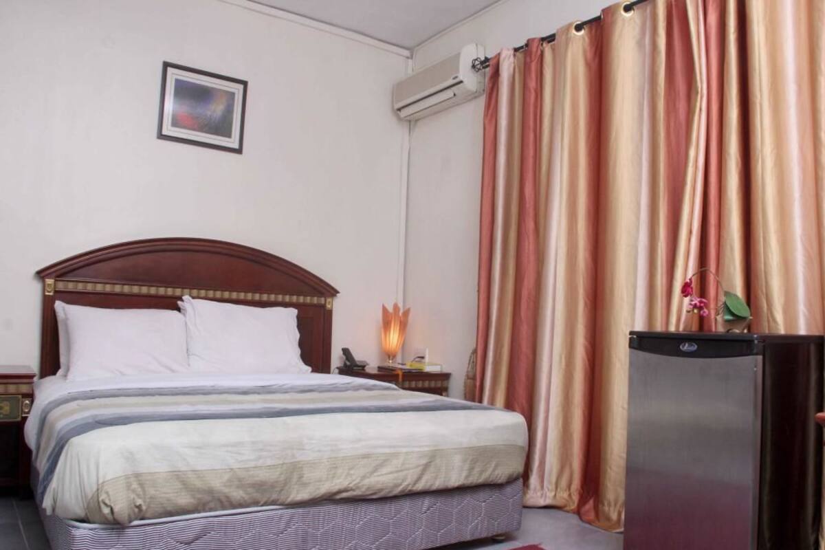 A hotel room with a bed, a nightstand, a lamp, curtains, and a refrigerator.