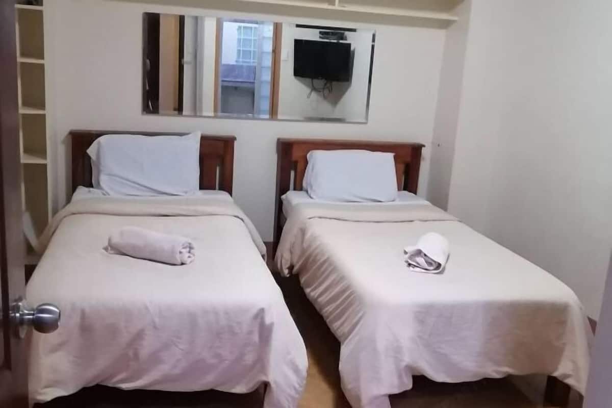 Two single beds with white linens and towels, a television mounted on the wall, and a window with a view of the outside.