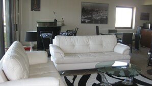 Una sala de estar moderna con un sofá blanco, una mesa de centro de vidrio y un tapete estampado de cebra. Hay una zona de comedor con una mesa y sillas, y una lámpara negra sobre una mesita auxiliar.