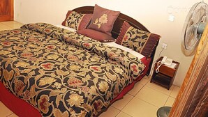A neatly made bed with a floral-patterned bedspread and matching pillows, a bedside table with a phone, and a standing fan. - Golden King Apartment Hotel (Lagos)