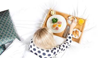 Une personne allongée sur un lit, avec un plateau contenant des aliments pour le petit-déjeuner, notamment des œufs, du jambon et des croissants, ainsi qu'un verre de jus d'orange.