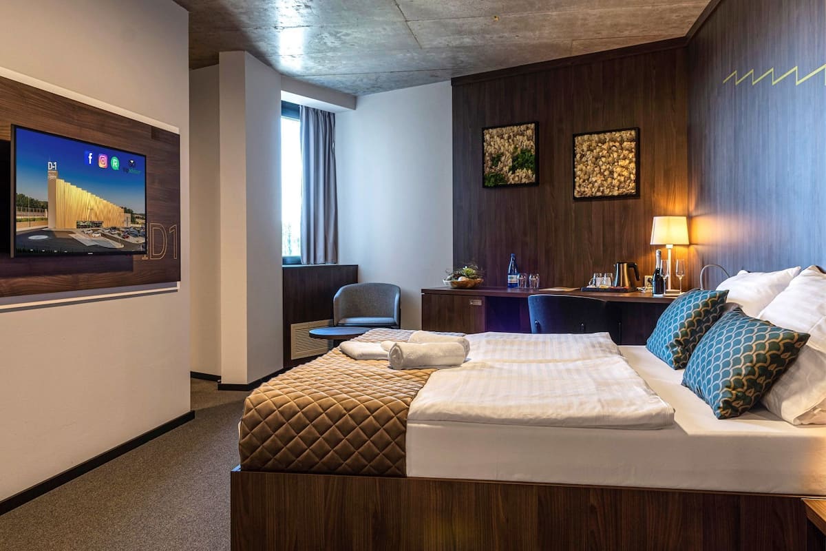Une chambre d’hôtel comprenant un lit, un bureau, une chaise, un téléviseur fixé au mur et des œuvres d’art accrochées au mur.