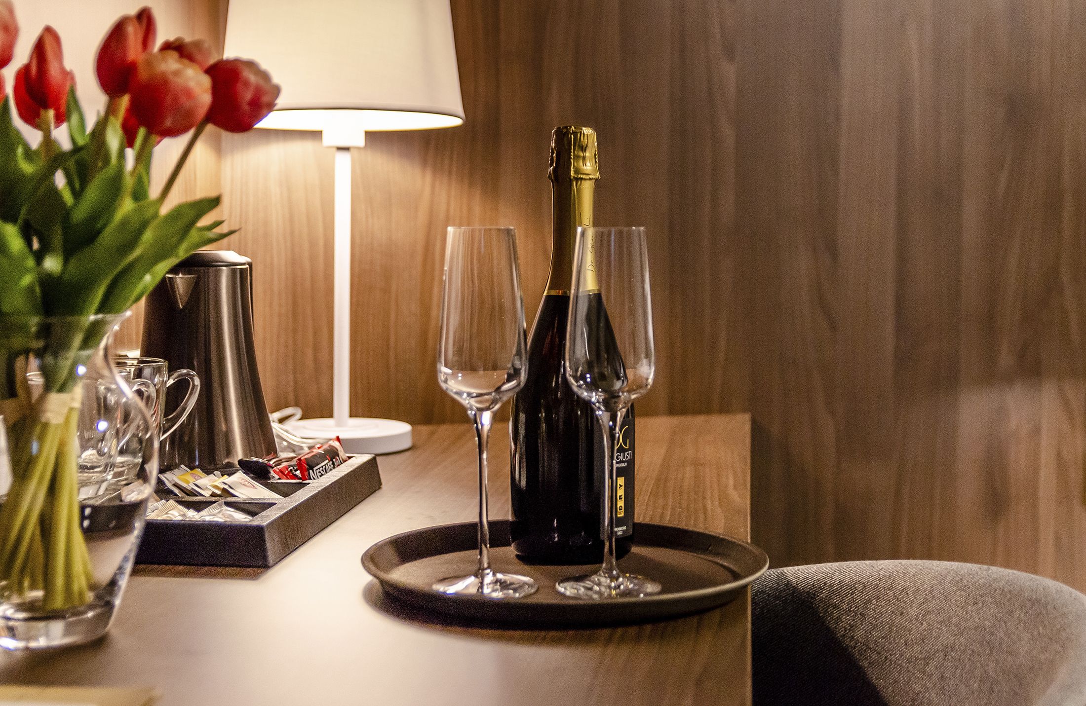 A table with a bottle of champagne, two glasses, a tray, a coffee maker, and a vase with red tulips.