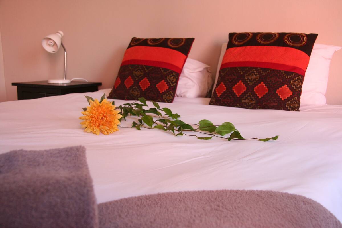 A neatly made bed with a single yellow flower and green leaves, two patterned pillows, a bedside lamp, and a dark nightstand.