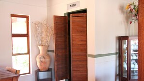 Une pièce avec une porte en bois portant l’inscription « Toilettes », un grand vase contenant des plantes séchées, une fenêtre et un meuble avec des portes en verre.