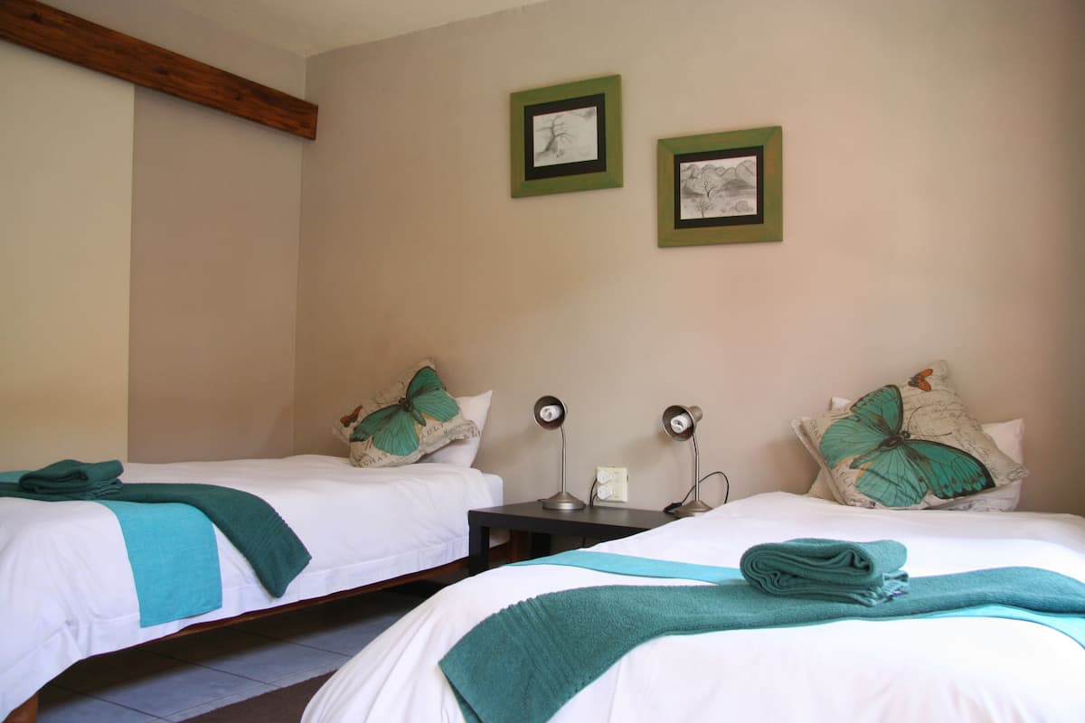 A hotel room with two beds, each with a green pillow and a teal blanket. There are two bedside tables with lamps.