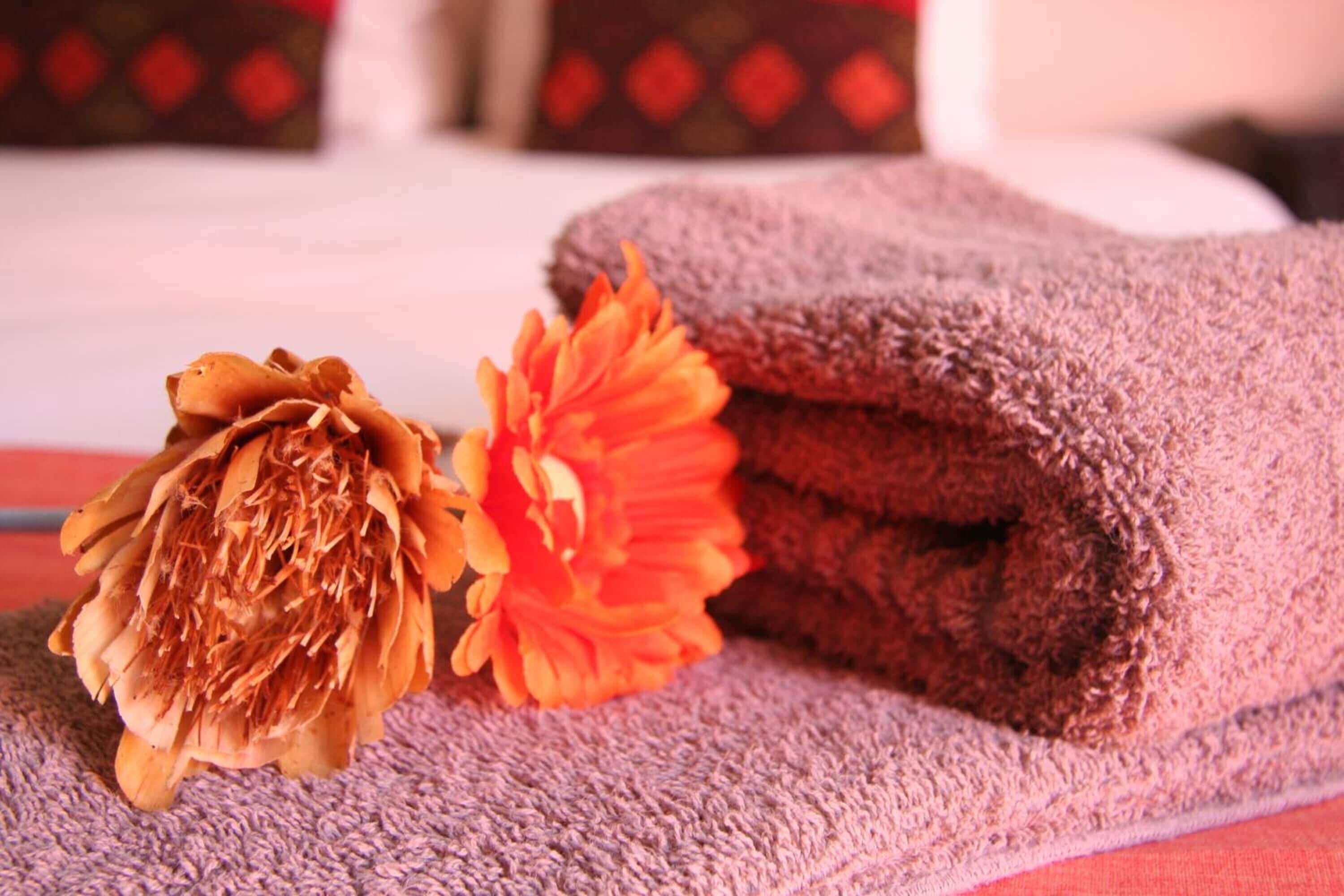 Ein ordentlich gefaltetes violettes Handtuch und eine getrocknete Orangeblüte auf einem Bett mit weißem Kopfteil und roten Kissen.