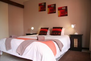 A neatly made bed with a white and orange bedspread, two pillows with red and brown patterns, and a brown throw blanket. There are two bedside tables with lamps, and three abstract paintings on the wall.